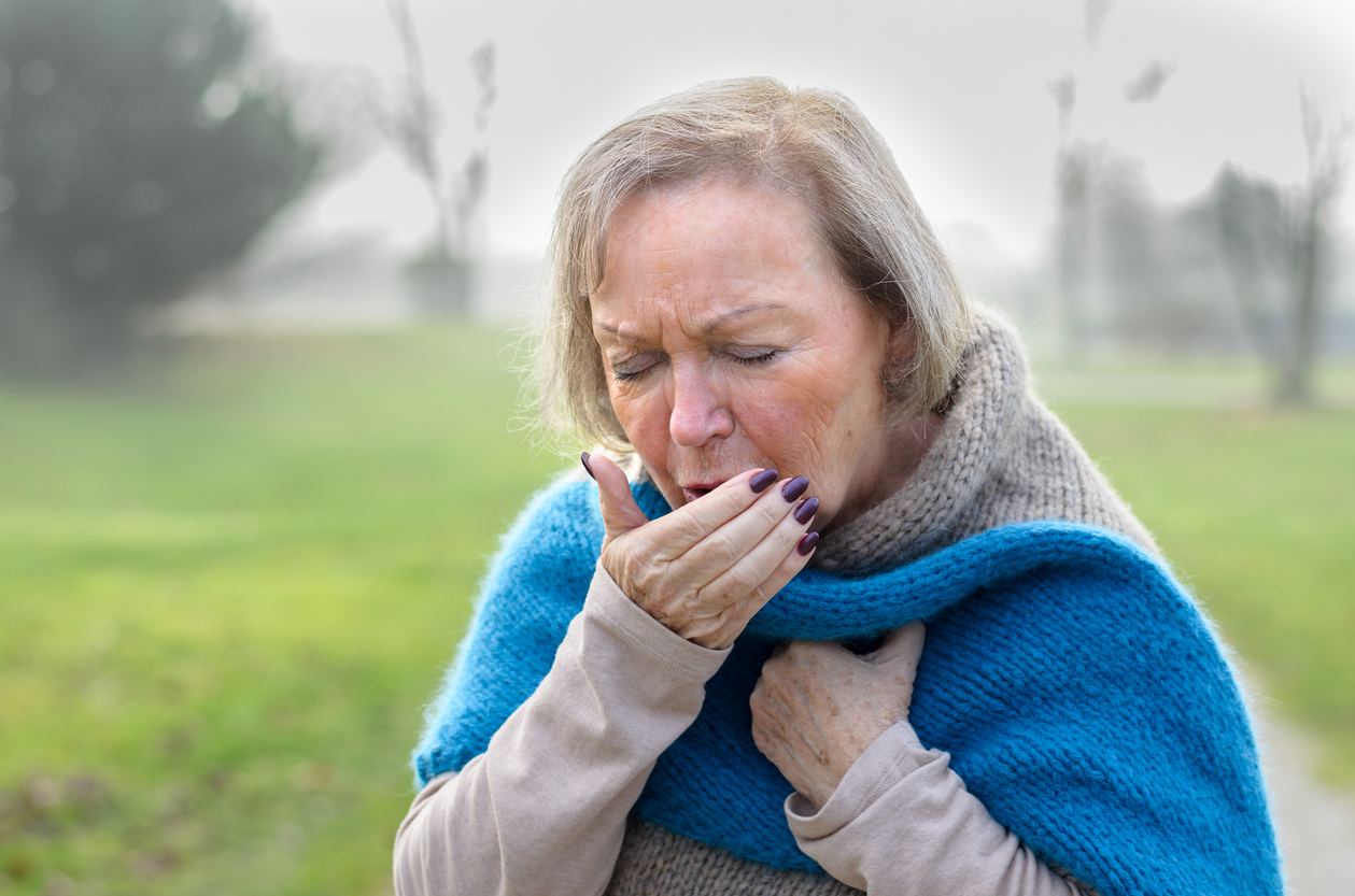 Elderly stylish attractive blond woman coughing or sneezing into her hand as she stands on a rural lane on a misty winter day